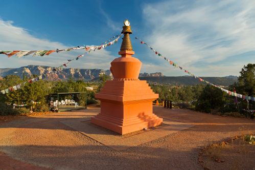A pink stupa surrounded by prayer flags, set against a backdrop of mountains and a clear blue sky.