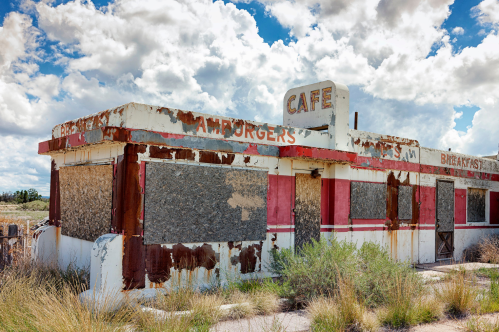 Abandoned diner with boarded windows, rusted exterior, and a sign reading "Cafe" under a cloudy sky.