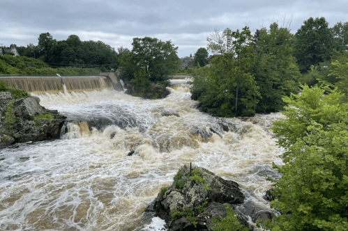 A rushing river cascades over a dam, surrounded by lush green trees and rocky outcrops under a cloudy sky.
