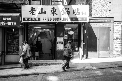 Black and white image of a fried dumpling shop with two people walking by on a city street.