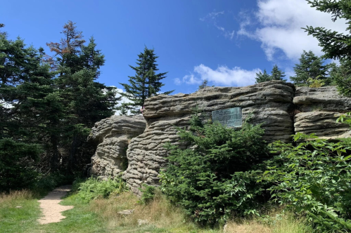 A rocky formation surrounded by trees, with a plaque mounted on the rocks, under a blue sky with scattered clouds.