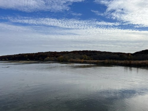 A calm river reflects the sky, with a wooded hillside in the background under a partly cloudy blue sky.