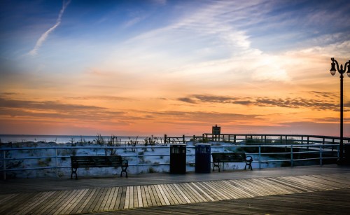 A serene beach boardwalk at sunset, with benches, trash cans, and a colorful sky reflecting on the horizon.