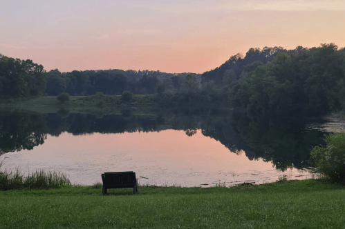 A serene lake at sunset, reflecting trees and a colorful sky, with a bench in the foreground on lush green grass.