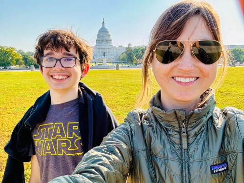 A woman and a boy smile for a selfie in front of the U.S. Capitol building on a sunny day.