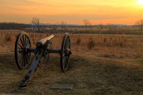 A historic cannon sits on grassy terrain at sunset, with a scenic landscape of trees and hills in the background.