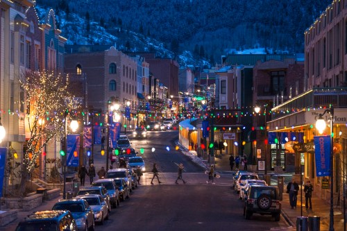A festive street scene at dusk, adorned with colorful lights and snow-capped mountains in the background.