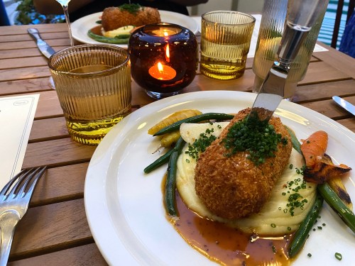 A plated meal featuring a crispy croquette on mashed potatoes, surrounded by green beans and carrots, with a candlelit ambiance.