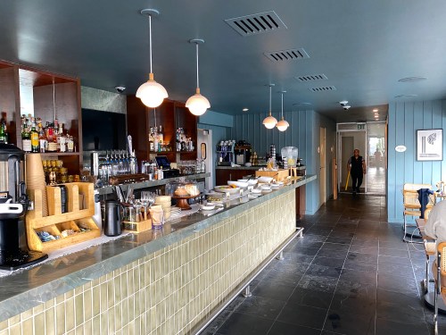 A modern bar area with a long counter, shelves of drinks, and a person walking in the background.