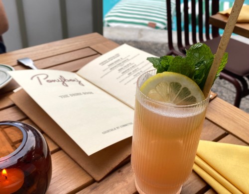 A refreshing cocktail with lemon and mint sits on a wooden table next to a menu, with a pool in the background.
