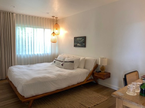 A cozy bedroom featuring a bed with white linens, a wooden nightstand, and large windows with sheer curtains.