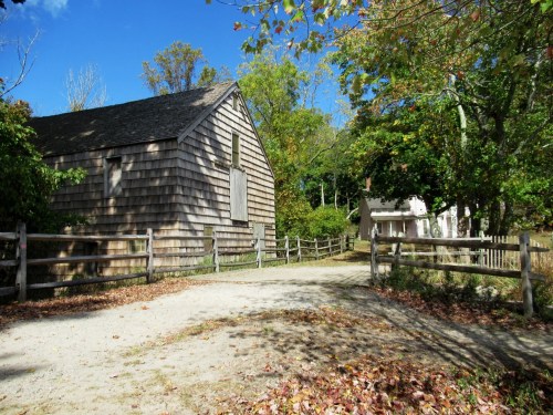 A rustic wooden building surrounded by trees and a dirt path, with a fence and another structure in the background.