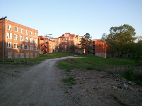 Abandoned brick buildings along a dirt road, surrounded by greenery and trees under a clear sky.