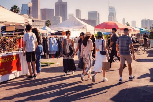 A bustling outdoor market with people walking among vendor tents and city skyline in the background.