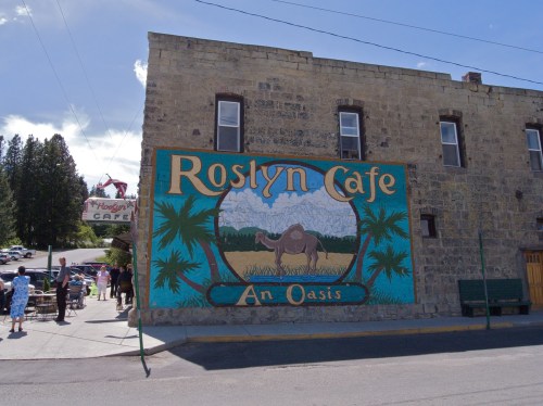 Mural of a camel and palm trees on the wall of Roslyn Cafe, with people dining outside on a sunny day.