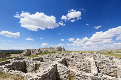 Ruins of ancient stone structures under a bright blue sky with fluffy clouds, surrounded by green grass.