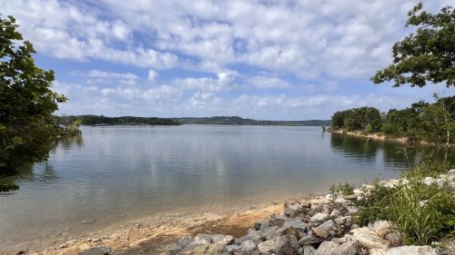 A serene lake view with calm waters, rocky shore, and lush greenery under a partly cloudy sky.