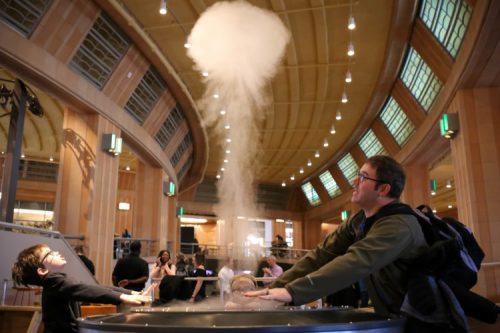 A man and a child interact with a steam vortex in a museum, surrounded by a spacious, well-lit interior.