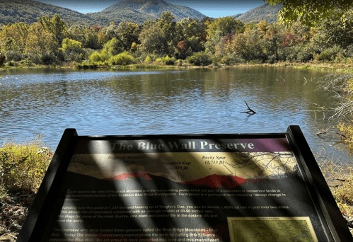 A scenic view of a lake surrounded by mountains, with a sign about the Blue Wall Preserve in the foreground.
