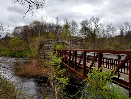 A rustic stone arch bridge over a river, surrounded by trees and cloudy skies.