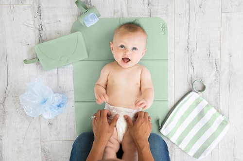 A smiling baby on a changing mat, with diaper supplies and a green striped bag nearby.