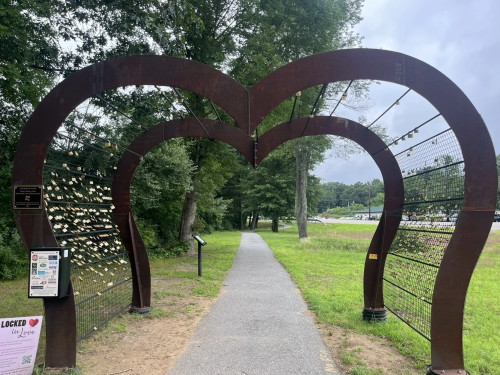 A heart-shaped archway with locks attached, leading to a path in a green park. Trees and a cloudy sky in the background.