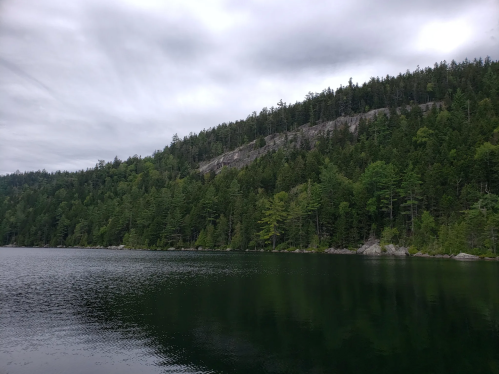 A serene lake surrounded by lush green trees and a rocky hillside under a cloudy sky.