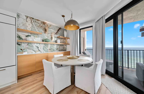 Modern dining area with a round table, white chairs, and a view of the ocean through large glass doors.