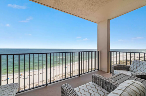 A balcony view overlooking a sandy beach and ocean under a clear blue sky, with outdoor seating.