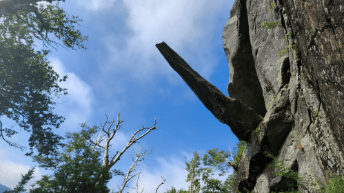 A tall, jagged rock formation juts out against a blue sky, surrounded by trees and wispy clouds.