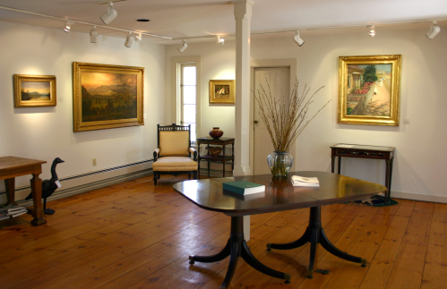 Art gallery interior featuring paintings on the walls, a wooden table, and a chair, with warm wooden flooring.
