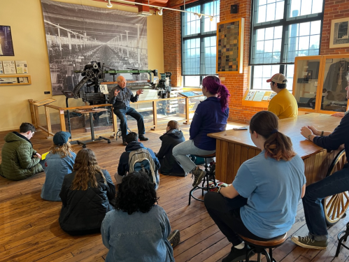 A group of people sits on the floor, listening to a speaker in a museum with industrial exhibits in the background.
