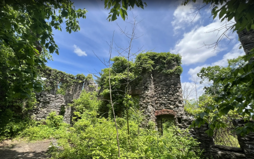 Ruins of a stone structure surrounded by lush greenery and trees under a bright blue sky with scattered clouds.