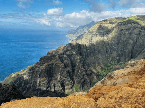 A panoramic view of rugged cliffs and lush greenery overlooking a blue ocean under a partly cloudy sky.