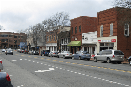 A street lined with shops and parked cars, featuring brick buildings and a cloudy sky.