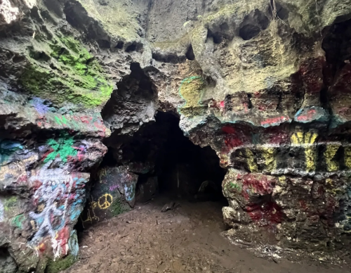 A colorful cave entrance with graffiti on the walls, surrounded by rugged rock formations and a muddy floor.