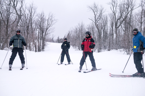 Four skiers stand on a snowy trail, surrounded by bare trees, wearing winter gear and ski equipment.