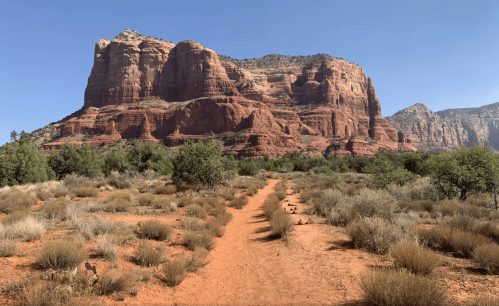 A sandy path leads through shrubs toward a large red rock formation under a clear blue sky.