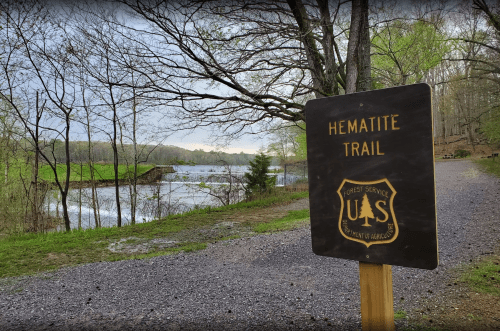 Sign for Hematite Trail near a river, surrounded by trees and a gravel path, with a cloudy sky above.