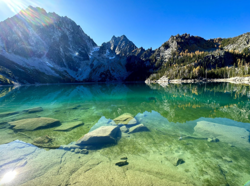 A serene mountain lake with clear turquoise water, surrounded by rocky peaks and lush forest under a bright blue sky.