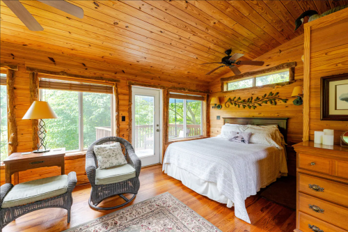 Cozy wooden bedroom with a queen bed, wicker chairs, and large windows overlooking greenery.