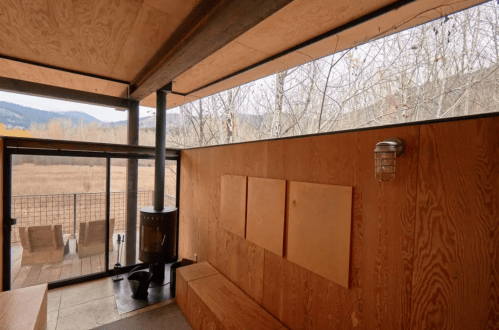 Interior of a modern cabin with wooden walls, a wood stove, and large windows overlooking a natural landscape.