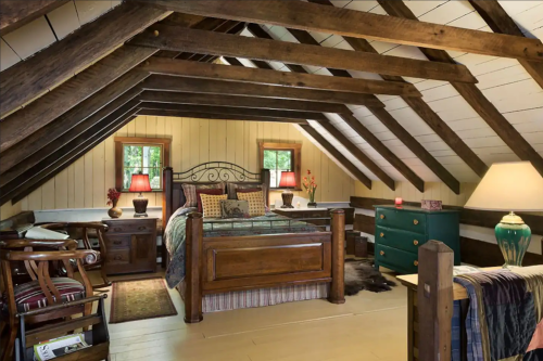 Cozy attic bedroom with wooden beams, a large bed, nightstands, and a green dresser, illuminated by warm lamps.