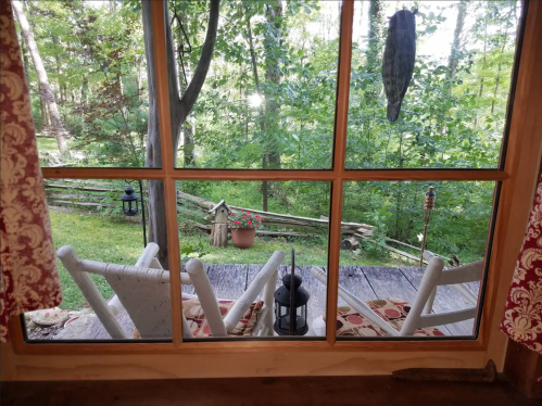 View through a window showing a wooden deck, rocking chairs, and a lush green garden with a flower pot.