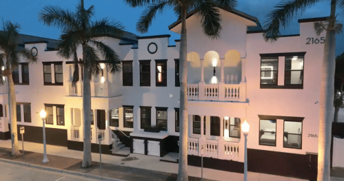 A modern two-story building with palm trees, featuring balconies and large windows, illuminated at dusk.
