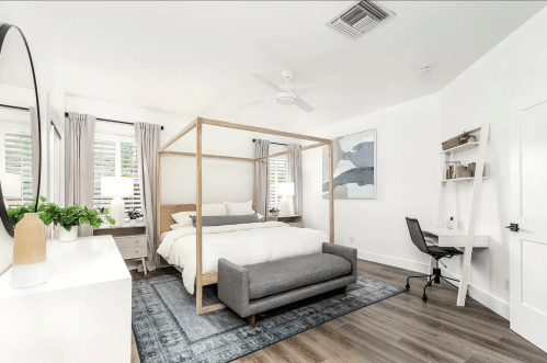A modern bedroom featuring a four-poster bed, a gray sofa, a desk, and light-colored decor with natural light.