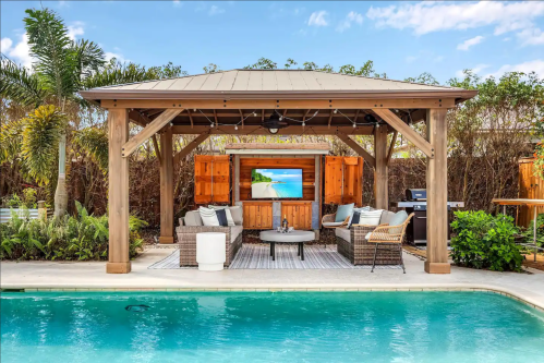 A cozy outdoor lounge area with a TV, surrounded by greenery and a pool, featuring comfortable seating and a grill.