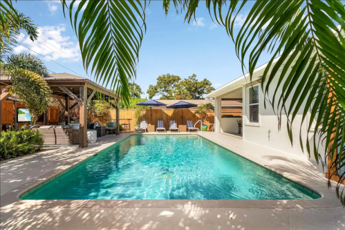 A serene backyard pool surrounded by palm trees, lounge chairs, and shaded areas under blue skies.