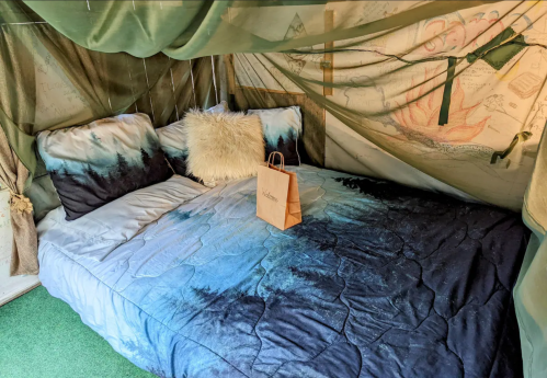A cozy tent interior with a bed featuring blue and white bedding, a fluffy pillow, and a small paper bag on top.