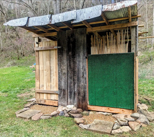 A rustic shed with a corrugated metal roof, wooden walls, and a green panel, set in a grassy area surrounded by trees.